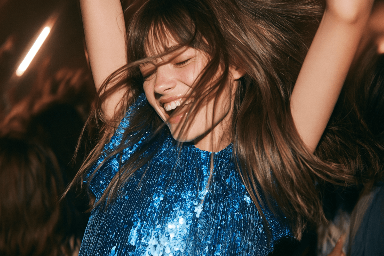 Close crop of a hand with chrome-painted nails lifting the edge of a blue satin dress away from the skin, the fabric falling in soft folds. A panel of bright blue disc sequins catches the light down the right of the frame.